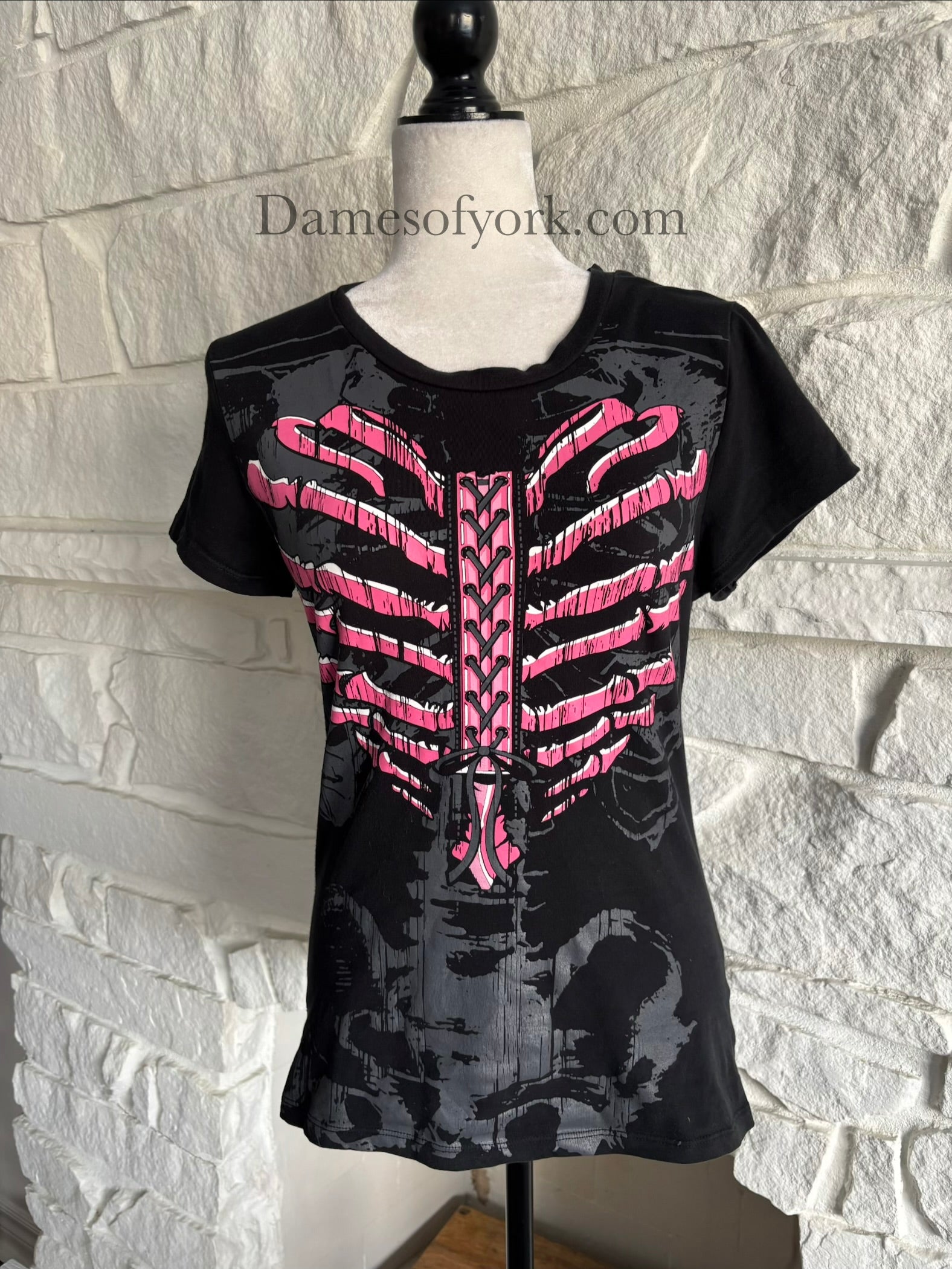 A close-up front view of a black gothic graphic t-shirt featuring a pink ribcage shaped like a heart with printed lace-up detail and a subtle grey skeleton design underneath, styled in an emo-inspired alternative design.