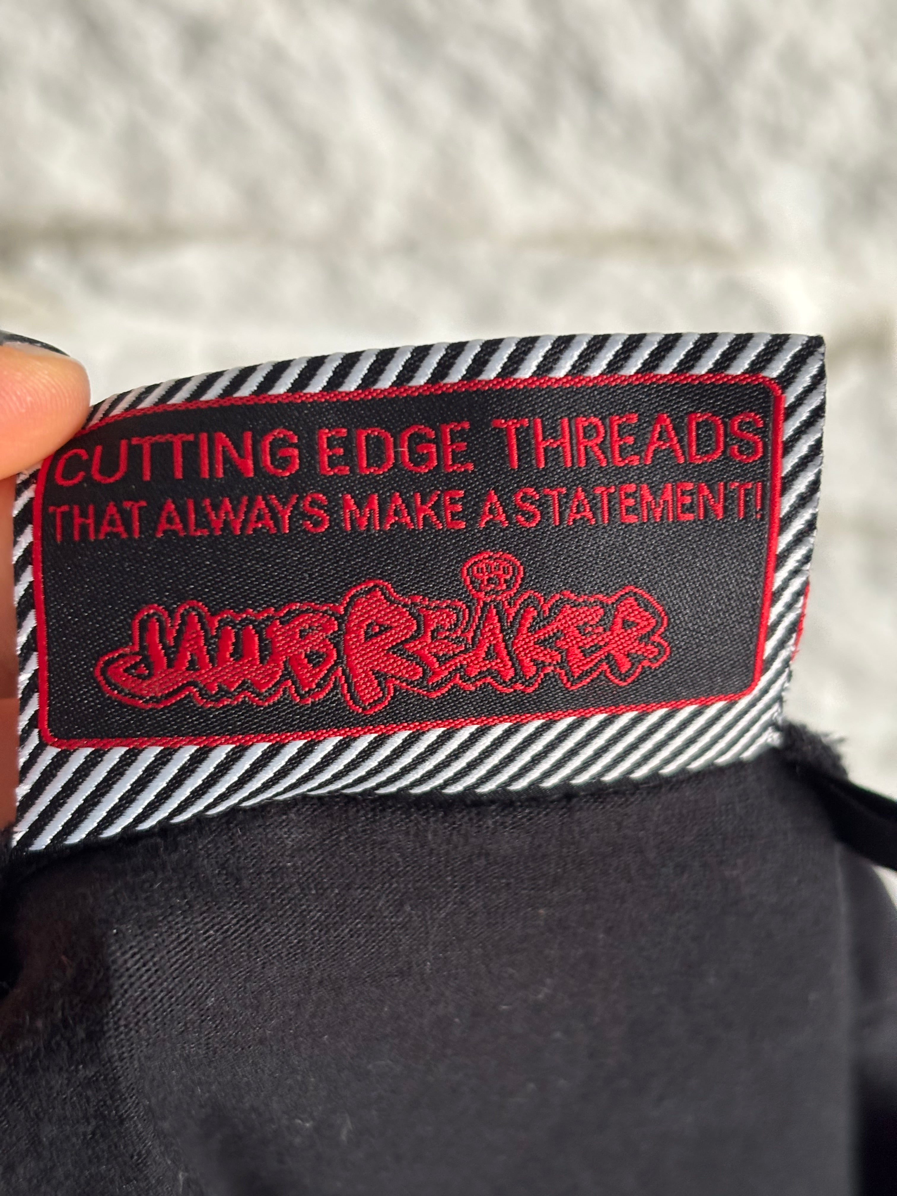 Textile label with red and white text on a black background, held against a textured fabric background. The text says "Cutting edge threads that always make a statement! Jawbreaker Clothing."