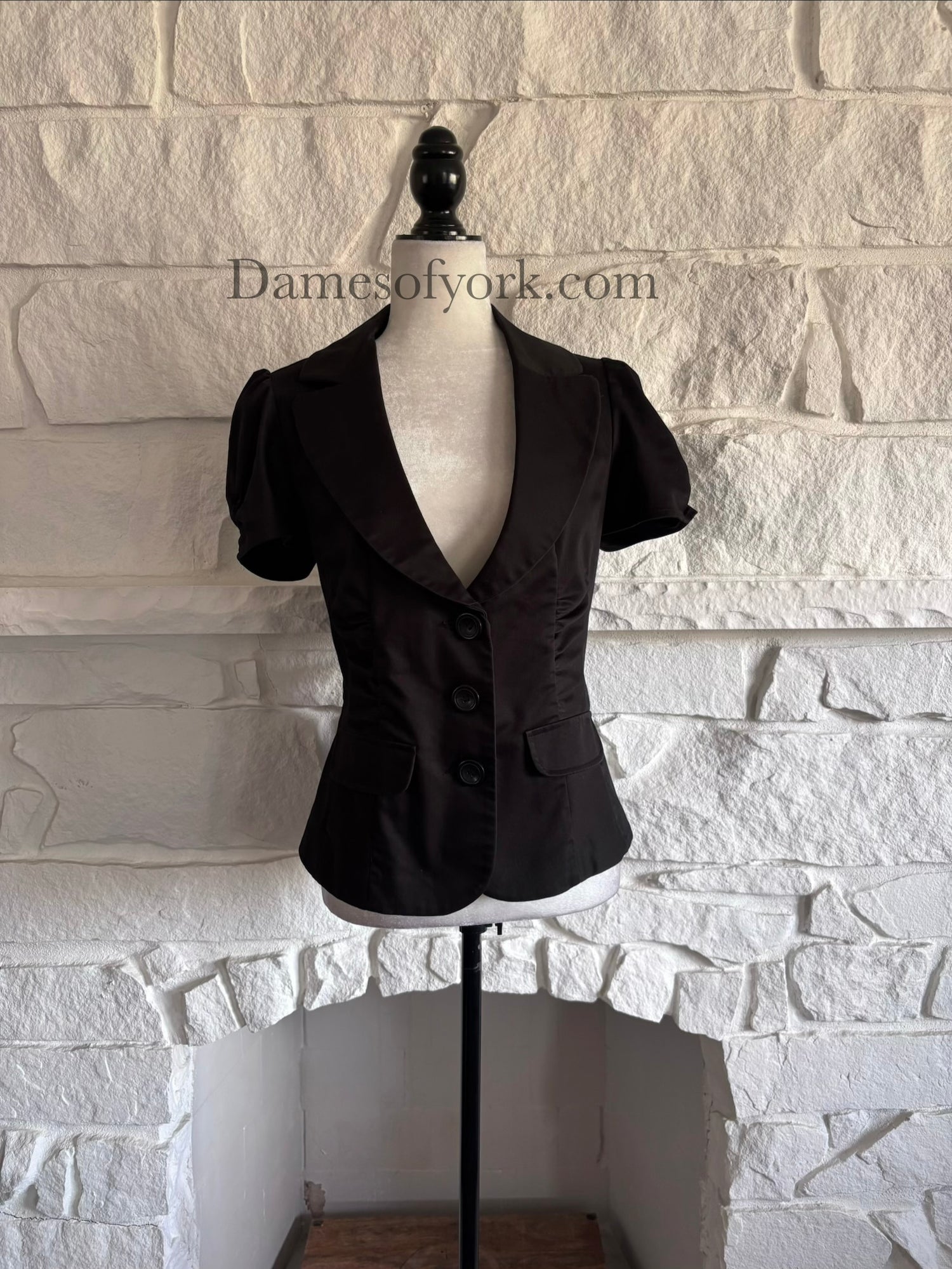 Front view of fitted black short sleeve blazer on mannequin with ruched waist and three-button closure