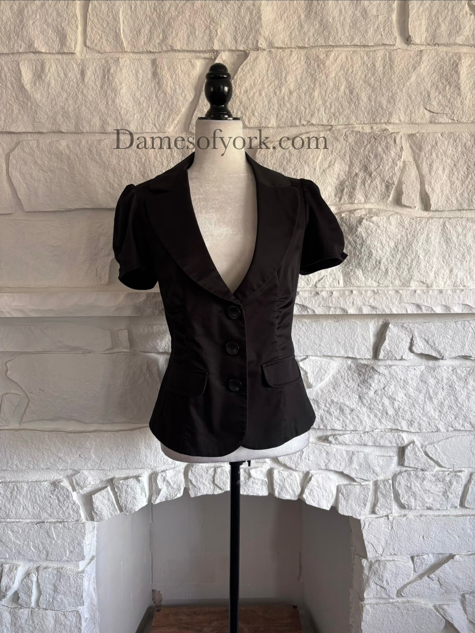 Front view of fitted black short sleeve blazer on mannequin with ruched waist and three-button closure