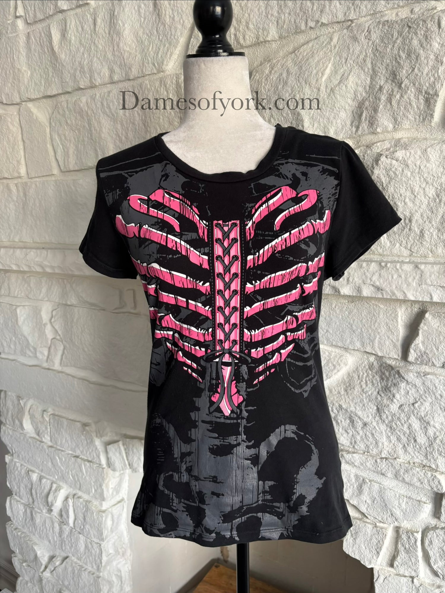 A close-up front view of a black gothic graphic t-shirt featuring a pink ribcage shaped like a heart with printed lace-up detail and a subtle grey skeleton design underneath, styled in an emo-inspired alternative design.